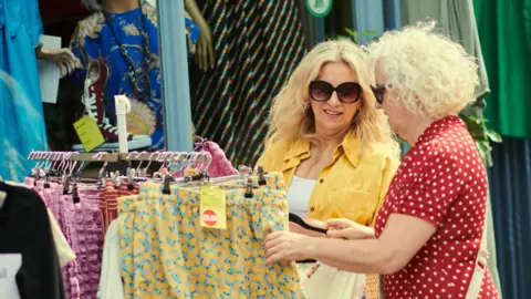 Two women in sunglasses shopping at an outdoor market, standing a rack of clothes including patterned yellow shorts.