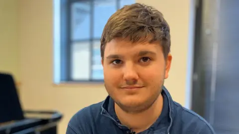 Alex Dunlop/BBC Mark Slaski has short brown hair and beard. He is wearing a navy blue shirt and is sitting in a room which has a piano on the left.