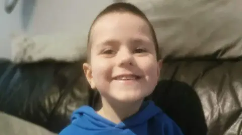 Family Handout Kyran Durnin - a young boy with short brown hair smiles at the camera while sitting on a dark leather sofa. He is wearing a blue hoodie.