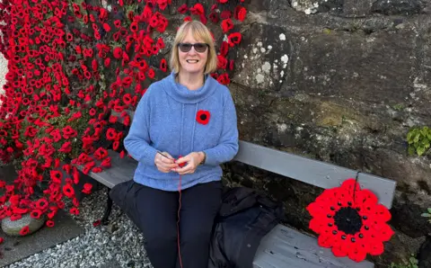 A woman sitting on a bench, wearing sunglasses, a blue jumper. She is surrounded by crocheted poppies