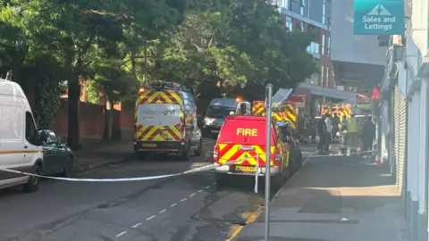 A picture of fire service and National Grid vans parked up outside the takeaway, with a cordon blocking off people from going anything further. There are several people stood outside the takeaway, to the right of the picture.