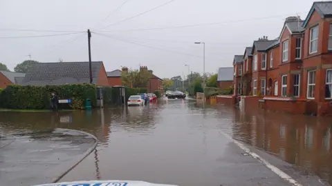 A wide view of a road that is submerged in brown flood water. There are houses on the right, where the flood water reaches, and there is a car in the distance turning away from the water