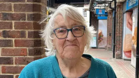 Phil Harrison/BBC An elderly woman with short, white hair and blue glasses. She is wearing a blue cardigan and standing in a shopping street near Greggs.