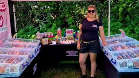 Supplied Samantha Khul wearing black shorts and a black T-shirt and black sunglasses. She is standing among three tables on which there are plastic boxes filled with sweets.