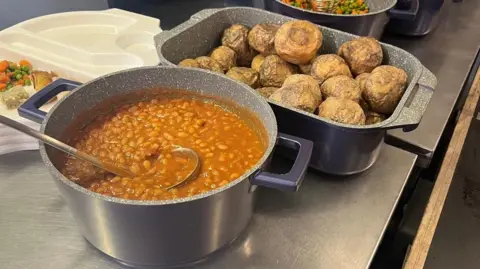 A large pot of baked beans with a serving ladel and a large oven tin full of baked potatoes.