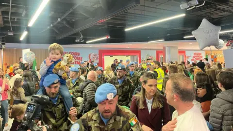 BBC A number of soldiers in camouflage clothing and blue hats walk through the airport. There are people with welcome home balloons and banners. 