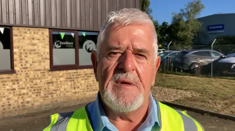 Luke Deal/BBC Mike Barrett wearing a hi-vis vest over a blue shirt. He has short grey hair and a goatee. He has his mouth slightly open and is standing in front of a brick building.