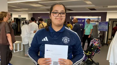 BBC/ Julia Bryson A young woman in a Manchester United track jacket holds up her results from a school reception area.