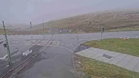 The Mountain Road at the Bungalow at the junction where the electric railway crosses the carriageway from Laxey Valley to go up Snaefell. Visibility is poor and the sky is grey, with the remains of the snow visible on the hillside opposite.