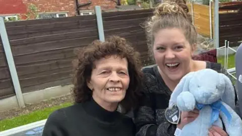 An older woman, two young women and a young man stand smiling at the camera as the young woman on the right takes a "selfie". The other young woman is holding a blue stuffed rabbit. 