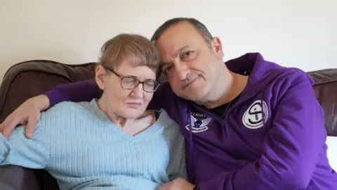 A man and a woman sitting on a brown leather sofa, and the man has his arm around the woman. She is wearing a blue top, has her eyes closed and is wearing glasses. The man is wearing a purple top and has short dark hair. 