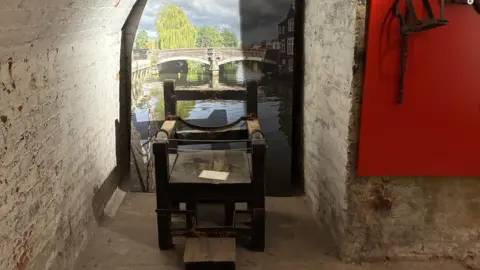 Emma Craig/BBC A heavy looking chair made of dark wood in Norwich Castle basement. It has arms and there is a metal bar towards the back of the chair. Behind it is a colourful photograph of the River Wensum in Norwich, with buildings on the right and a bridge in the distance.