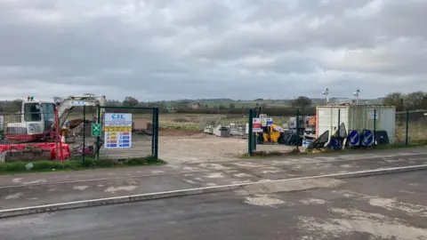 Daniel Mumby Picture shows fenced land with green scenery in the background and from within the fences there are road signs, a storage unit and a digger.