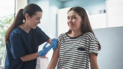 A woman is getting a flu vaccine from a nurse who is wearing a navy uniform and blue gloves. The background is blurred and shows a health clinic with plants outside the window.