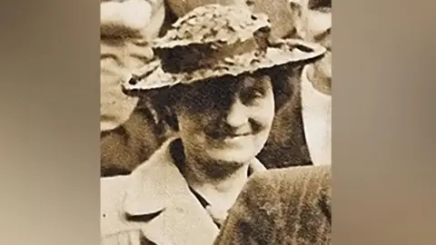 Avon and Somerset Police A sepia tone picture of Louisa Dunne. She is wearing a collared coat and wide brimmed hat. She is smiling at the camera in a crowd of people. 