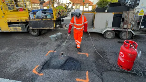 Simon Dedman/BBC Man in orange high viz with a helmet repairing a pothole. He is using a gas powered heater to work on smoothing the pothole before filling it with material.