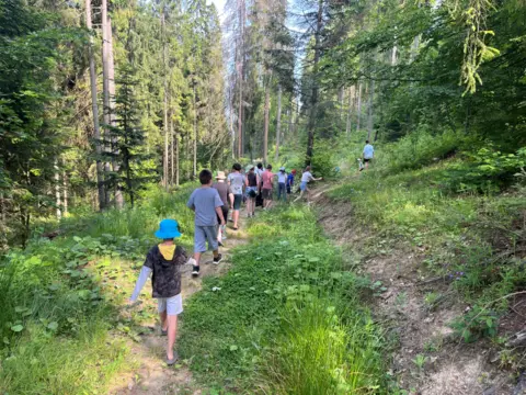 Children walk in a line on a hiking path surrounded by trees