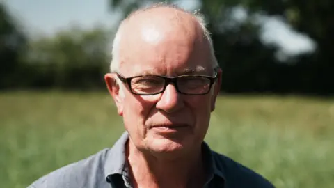 John Fairhall/BBC John Pawsey, who has thin white hair around the side of his head, stands in a field. He wears glasses and a dark grey shirt. He is squinting slightly due to the sun.