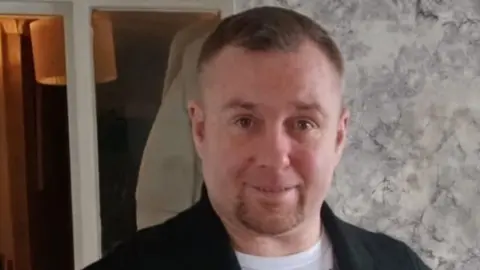 A man with short brown hair and a short goatee beard wears a black jacket and white t-shirt.