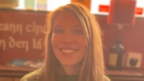 Maureen has long brown hair and is wearing a green jumper. She is sitting in a pub with red walls and signs in Irish behind her, also some bottles on display. 