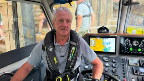 An older man with short white hair looking at the camera. He is wearing a grey polo shirt and a black life jacket. The controls of the boat are behind him.