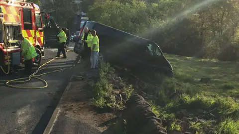 Chris Procter A black transit style van half in a field half on the pavement. behind it is a fire truck and a flipped over car as well as various police and fire officers.