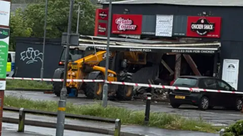The yellow JCB crashed into the damaged building. The black building has no window, with rubble inside. Police tape surrounds the premises. 