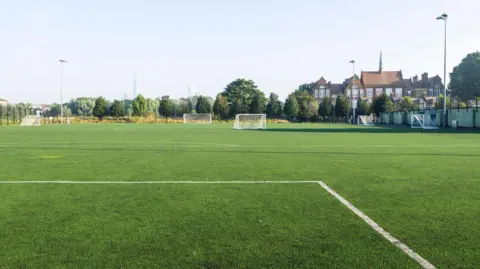 The image shows an empty leisure field in a residential district, with a view of the city in the distance. The sky is cloudless and light blue. The white lines of a football pitch can be seen in the foreground of the image.