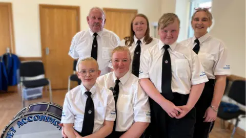 Evie sitting on her mum's knee with her two sisters, aunt and grandfather standing smiling beside a drum.