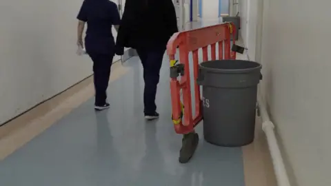 Two people are walking down a corridor with a blue floor and near the wall on the left is a large orange barrier with a large brown bucket inside. 