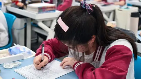 A teenager sits at a desk with her head down looking at a paper. She is writing on it with black pen. Stationery and a teddy bear sits on her desk. In the background are other teenagers at desks, all spaced out, in a classroom.