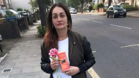 Met Police Photo of a petite woman with long dark hair carrying a flower and a book, wearing a white top underneath a dark jacket standing on a pavement. She has hoop earrings and looks directly at the camera.