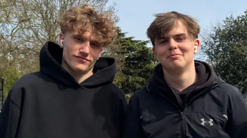 Two teenage boys in black jumpers look at the camera. 
