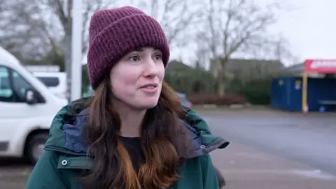 A woman with brown hair purple wooly hat and green jacket looking off camera