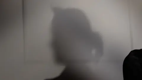 The shadow of Emma’s head and shoulders is cast against a pale wall. Her face and identifying features are not visible.