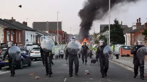 Getty Images Four riot police, holding shields walk down a residential street in Southport on a summer's evening. Bricks are strewn over the road, and in the background, a fire is burning. Behind the four police in the foreground, a line of police with longer shields follow them. 