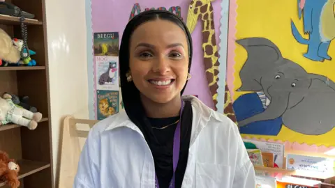 Teacher Fahima Miah smiling at the camera with a white shirt and black top underneath. She is wearing a hijab and has children's art behind her which shows an elephant and a giraffe