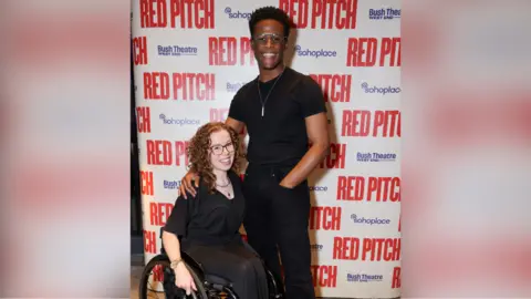 Getty Images Amy Trigg and Jordan Benjamin. Both are wearing black and smiling at the camera. They are standing in front of a promotional board at the Soho Place theatre in London.