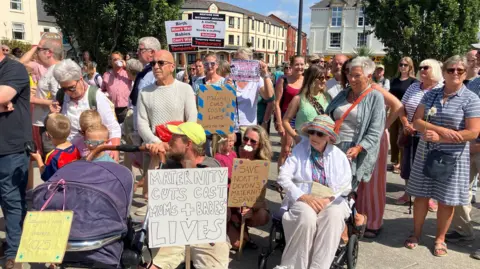 A crowd gathers in a town square during a protest to save maternity services. People of different ages hold placards reading messages such as “Maternity cuts cost mums + babies lives” and “Save North Devon’s maternity services,” while others stand with prams and wheelchairs in bright sunshine.
