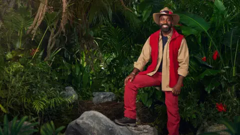 ITV Melvin Odoom wearing his jungle outfit, standing with his foot on a rock in a jungle scene 