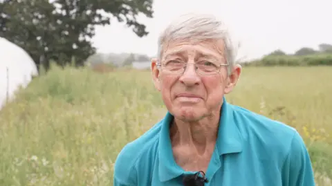 A man with short white hair is standing in a grassy field. He is wearing a teal, collared shirt with a small black microphone clipped to it. In the background, there are tall grasses, trees, and a white greenhouse. The sky is overcast.