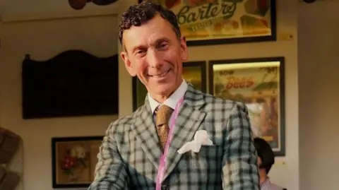  Matthew Upham looking at the camera. He is wearing a grey checked jacket, a light brown tie and a pink lanyard