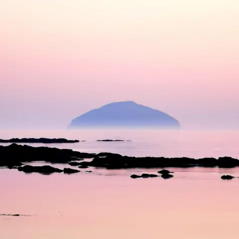 Jane Reith Soft pastel hued blend in the sky, reflecting on calm waters with a distant, misty island silhouetted against the horizon.