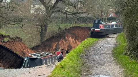 Shropshire Fire and Rescue Service Two canal boats can just about be seen in a drained section of canal. Another boat can also be seen teetering over the edge of the hole.