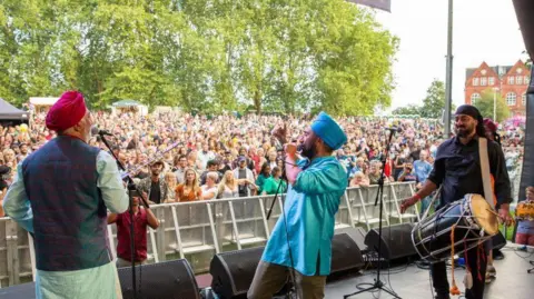 A band plays on a stage in the middle of a park. The photo is taken from the stage and there's a crowd of people standing looking towards the stage.