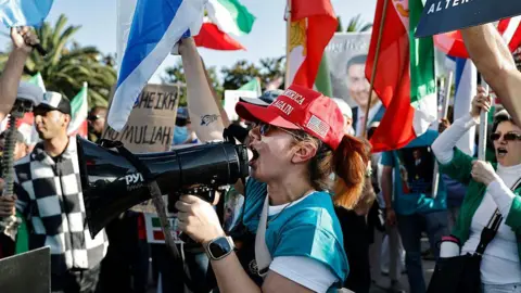 A woman in a Make America Great Again hat chants for regime change in Iran into a bullhorn during a protest in West LA in support of President Trump. A crowd behind her waves US, Israeli and Iranian flags. 