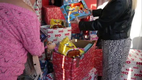 Cuboid Two women in a shipping container looking through carboard boxes full of gifts. Two spongebob squarepants toys are visible in the red and white boxes