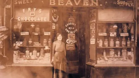 Colin Morris Colin's great aunt standing in front of a sweet shop that was later turned into the newsagents. The image is very old and in sepia tones.