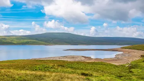 BBC Weather Watchers/ Murray McLaren The reservoir which is a large body of water surrounded by green hills. A coarse beach can be seen to the right of the photo in front of the water. The sky is light blue with large clouds.