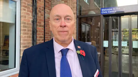 Councillor Rob Wharton is standing outside a building, wearing a blue suit jacket, pink shirt and blue spotted tie. A sign above a steel and glass door says "Welcome to the Civic Centre".